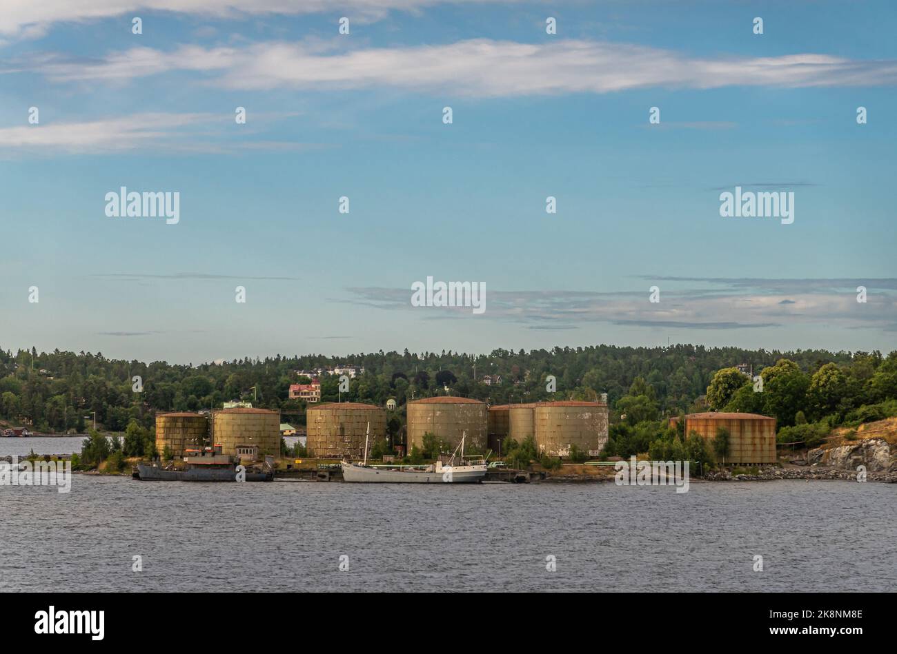 Schweden, Stockholm - 16. Juli 2022: Der alte Öl- und Gashafen auf der Insel Stora Hoggarn zeigt bräunte zylindrische Tanks und einen Pier mit 2 älteren Schiffen unter b Stockfoto