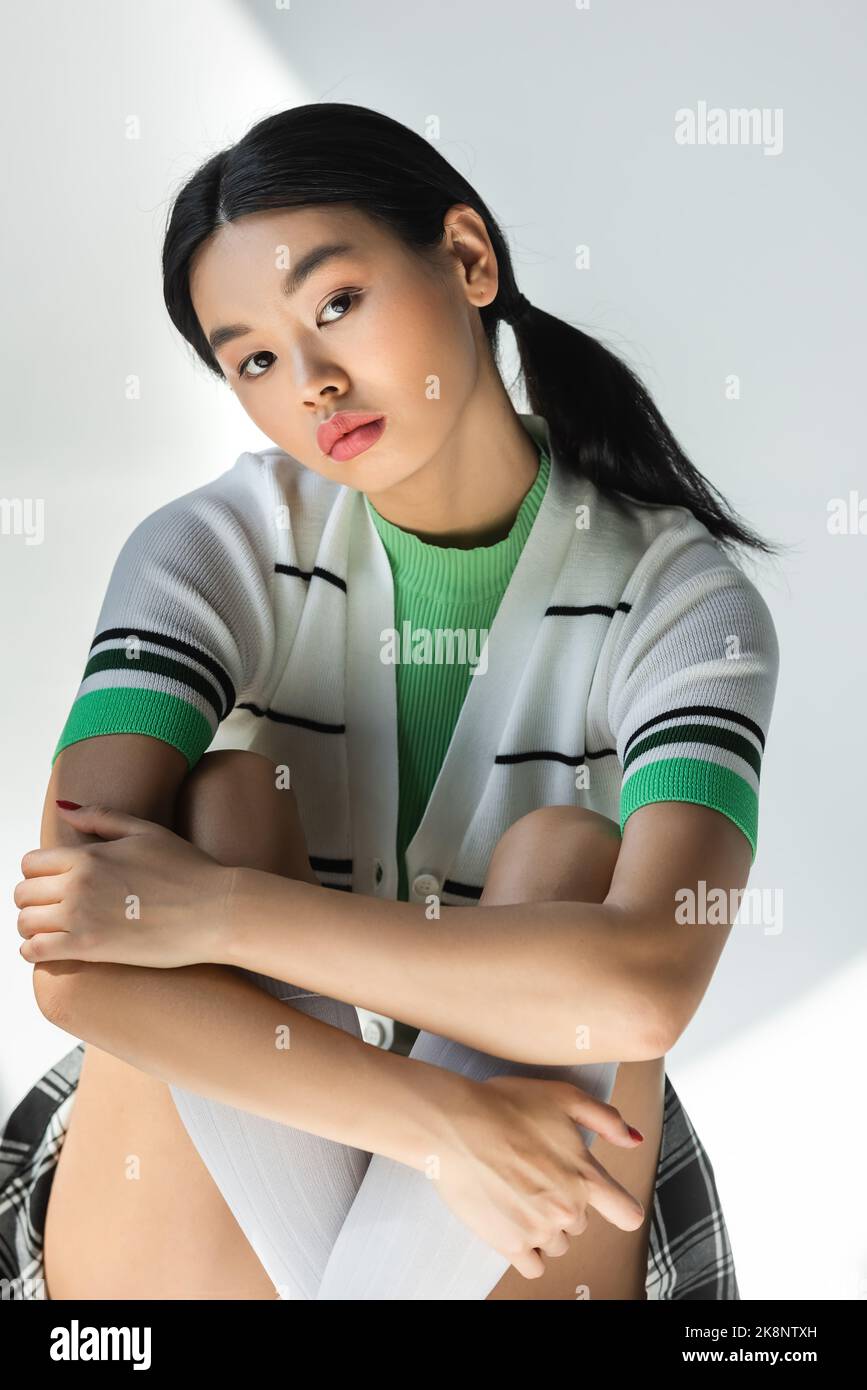 Junge asiatische Frau in gestreifter Strickjacke umarmende Knie auf grauem Hintergrund Stockfoto