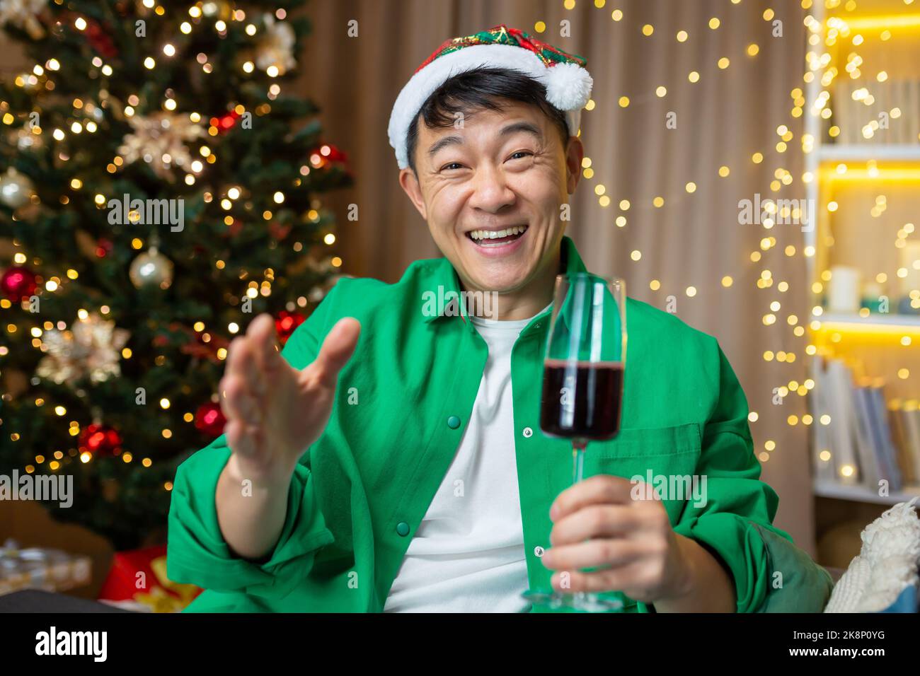 Fröhlicher asiatischer Mann an weihnachten und Neujahr begrüßt Freunde, Mann spricht bei einem Videoanruf mit Kollegen, die zu Hause auf dem Sofa sitzen, mit Hut und einem Glas Wein, Blick auf das Internet, Blick auf die Kamera und lächelnd. Stockfoto