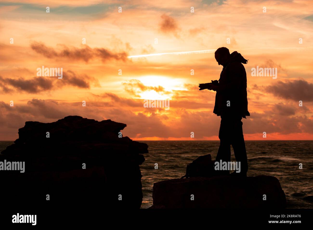 Portland, Dorset, Großbritannien. 24. Oktober 2022. Ein Fotograf trotzt dem wehenden Meereswind, um den Sonnenuntergang in Portland Bill, Dorset, an einem für die Saison unangenehmen Tag im Süden Englands einzufangen. Kredit: Peter Lopeman/Alamy Live Nachrichten Stockfoto