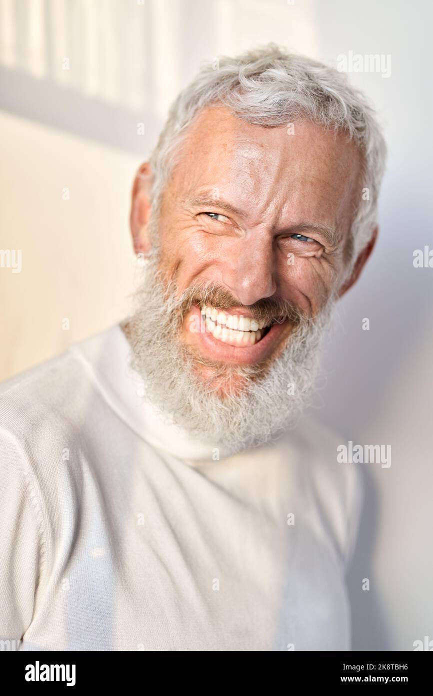 Lächelnder bärtiger älterer Mann, der lachend an der weißen Wand im Sonnenlicht steht. Stockfoto