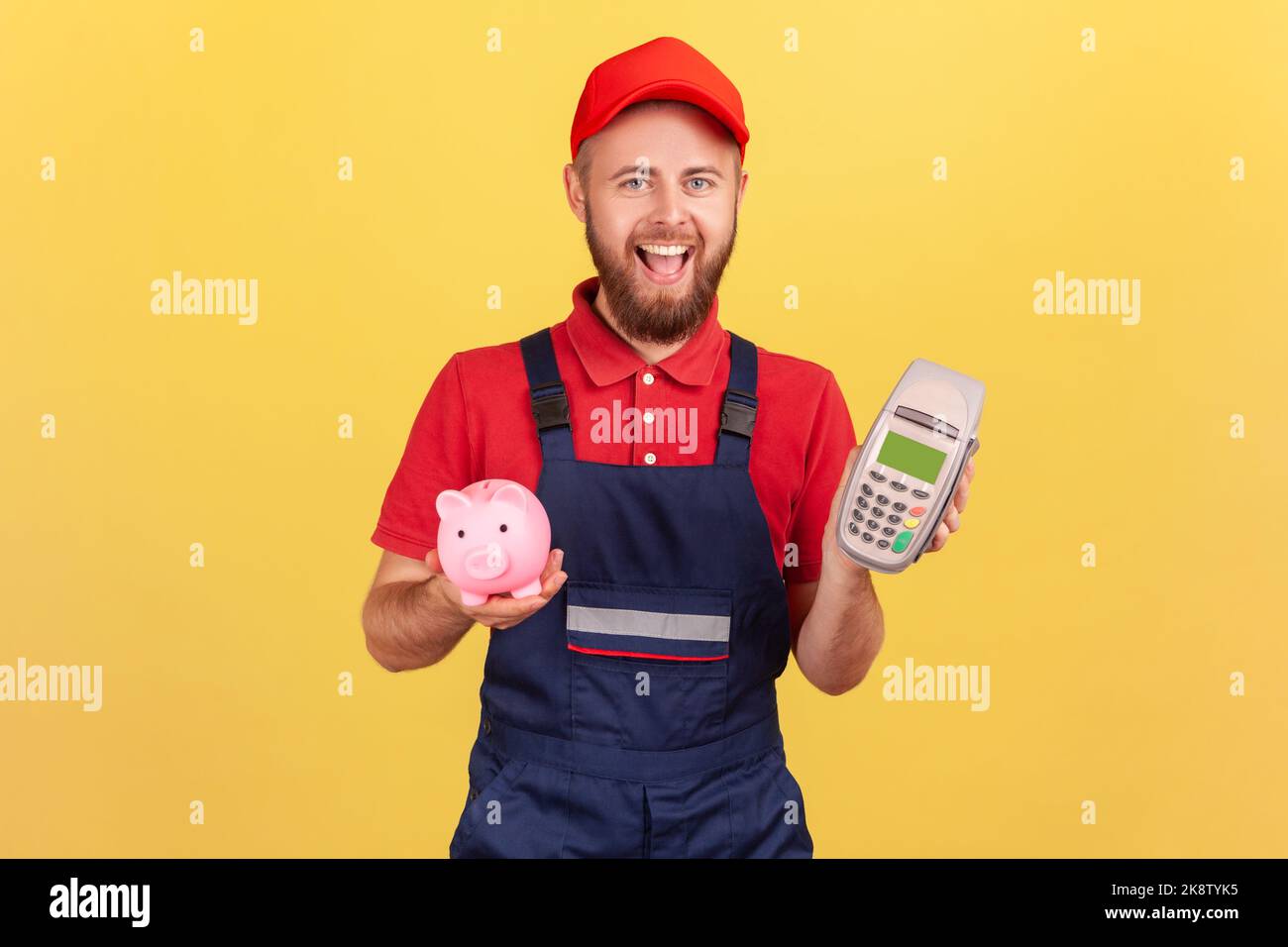 Fröhlicher bärtiger Arbeiter Mann hält in der Hand POS kontaktloses Zahlungsterminal und Sparschwein, Cashback, mit paypass für Service-Zahlung. Innenaufnahme des Studios isoliert auf gelbem Hintergrund. Stockfoto