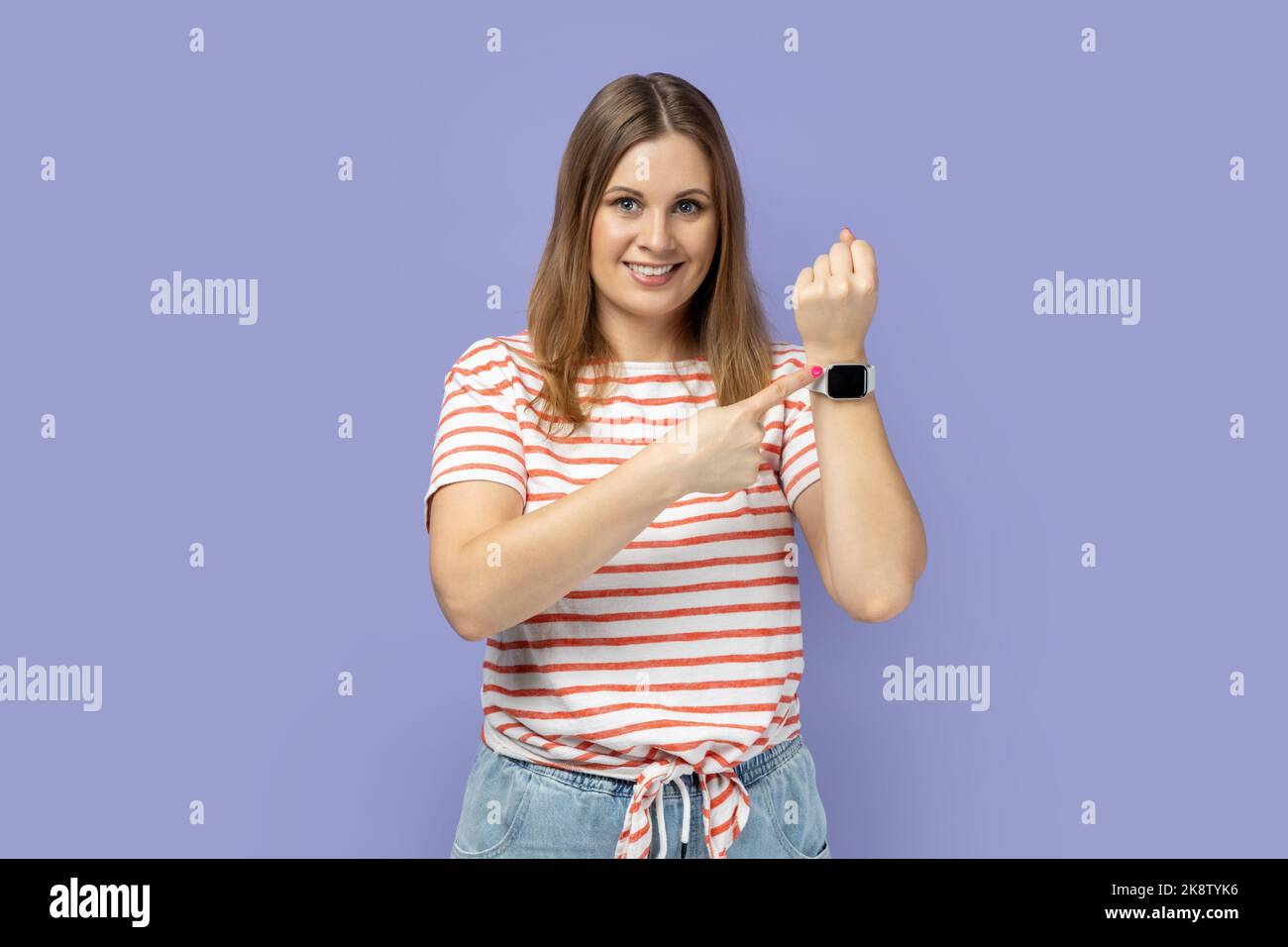 Gutes Ergebnis und pünktlich. Porträt einer blonden Frau, die im Stehen ein gestreiftes T-Shirt trägt, seine smarte Uhr zeigt und mit einem dutzenden Lächeln auf die Kamera blickt. Innenaufnahme des Studios isoliert auf violettem Hintergrund. Stockfoto