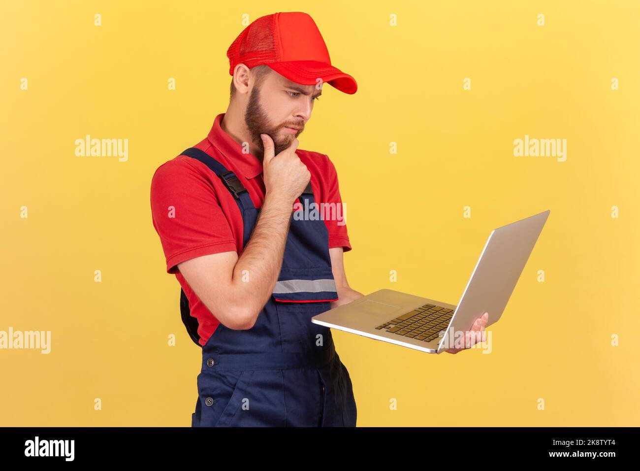 Seitenansicht eines nachdenklichen, nachdenklichen Arbeiters, der auf einem Laptop steht und arbeitet, tief in Gedanken ist, das Kinn hält, blaue Uniform und rote Mütze trägt. Innenaufnahme des Studios isoliert auf gelbem Hintergrund. Stockfoto