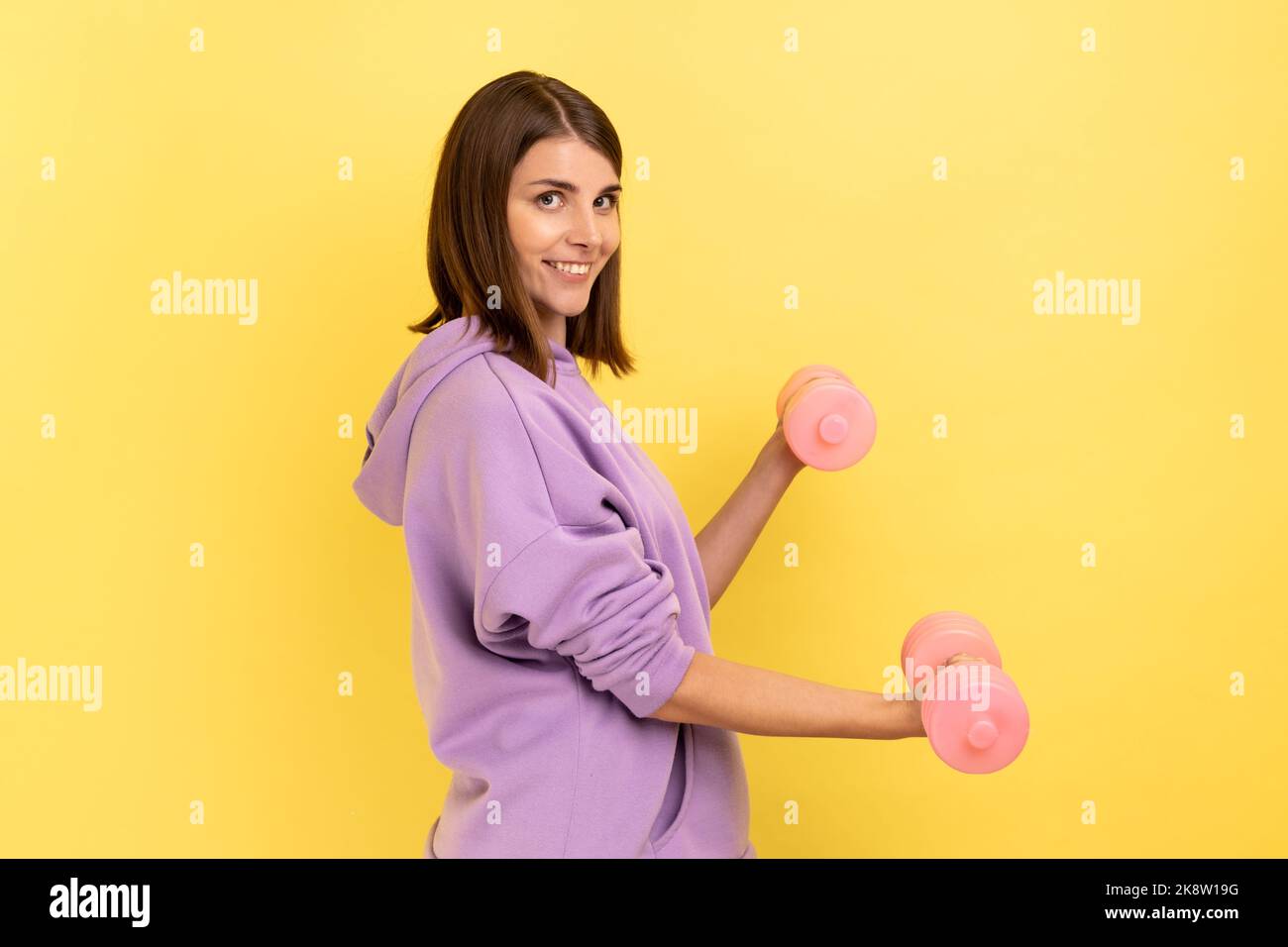 Seitenansicht einer Frau, die mit rosa Hanteln Fitness-Übungen macht, Hände trainiert, mit einem Lächeln auf die Kamera blickt und einen violetten Hoodie trägt. Innenaufnahme des Studios isoliert auf gelbem Hintergrund. Stockfoto