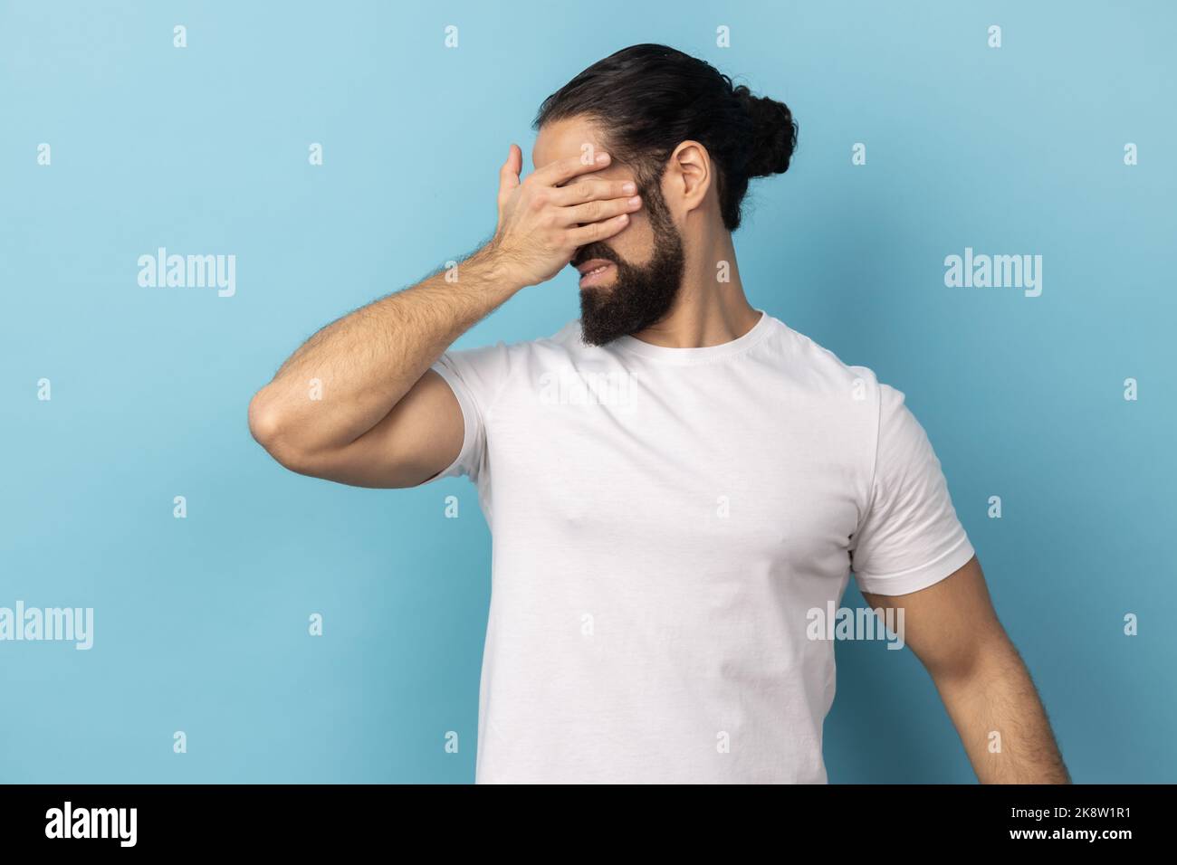 Ich möchte das nicht ansehen. Porträt eines verängstigten Mannes mit Bart, das weißes T-Shirt trägt, die Augen mit der Hand bedeckt und sich weigert, zuzusehen. Innenaufnahme des Studios isoliert auf blauem Hintergrund. Stockfoto