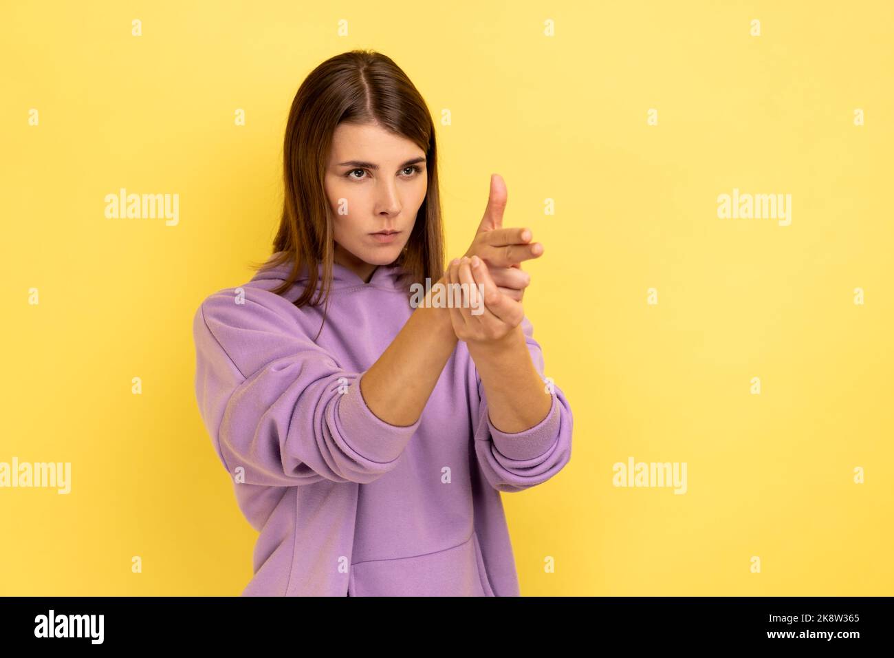Ich werde Euch töten. Porträt einer wütenden Frau, die mit Fingergewehren auf die Kamera zeigt, mit Schießdrohungen droht, Hände imitieren Waffen und trägt einen violetten Hoodie. Innenaufnahme des Studios isoliert auf gelbem Hintergrund. Stockfoto