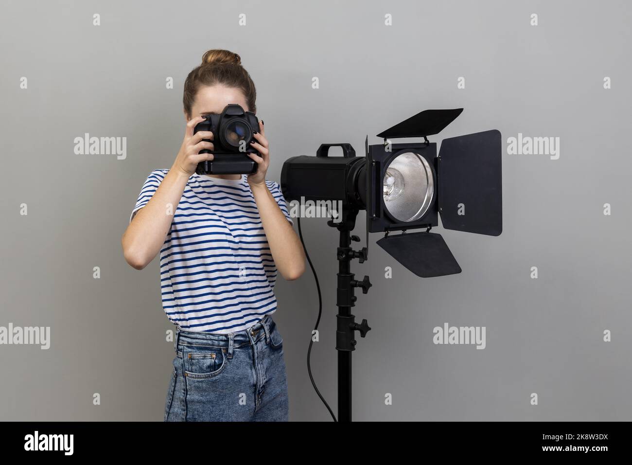 Porträt einer fleißigen Fotografin mit gestreiftem T-Shirt, die im Fotostudio fotografiert und dabei Fotozellen und Reflektoren verwendet. Innenaufnahme des Studios isoliert auf grauem Hintergrund. Stockfoto