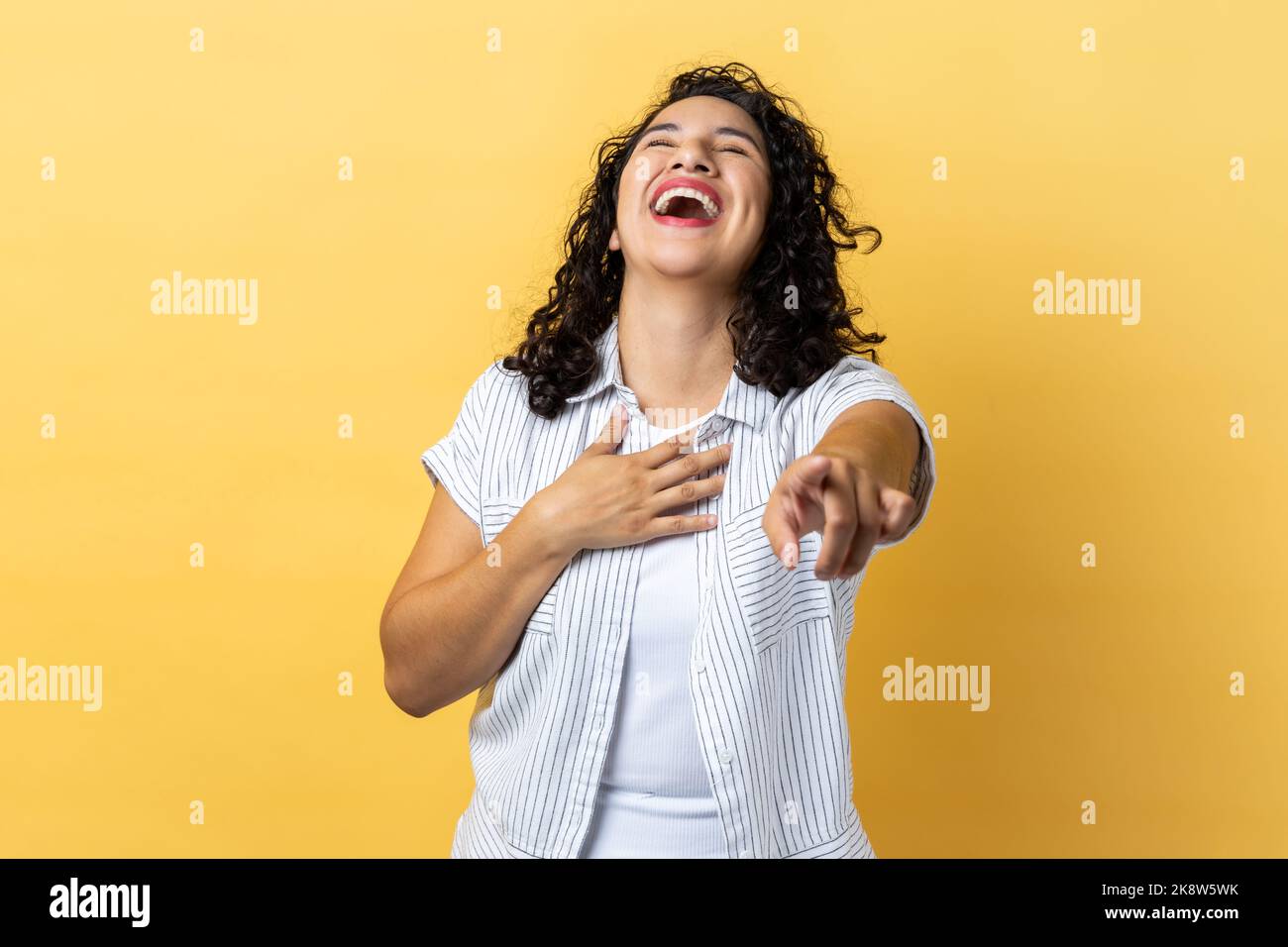 Porträt einer glücklichen Frau mit dunklen welligen Haaren, die laut lacht und mit dem Finger auf die Kamera zeigt, lustigen Witz hört, den Bauch hält. Innenaufnahme des Studios isoliert auf gelbem Hintergrund. Stockfoto