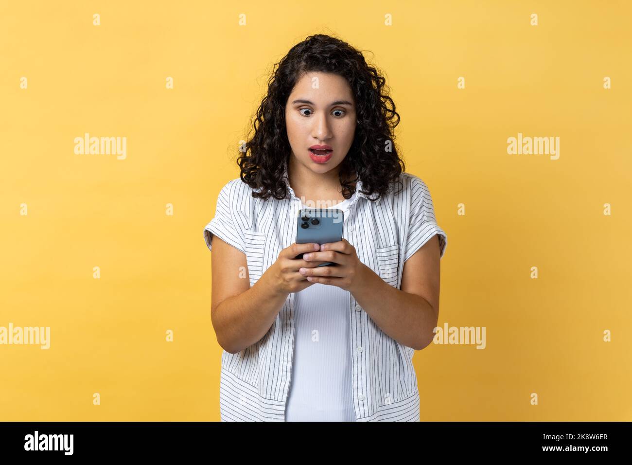 Porträt einer schockierten, staunenden Frau mit dunklem, welligen Haar, die Mobiltelefon benutzt und schockierende Nachrichten in sozialen Nachrichten liest und mit großen Augen auf den Bildschirm blickt. Innenaufnahme des Studios isoliert auf gelbem Hintergrund. Stockfoto