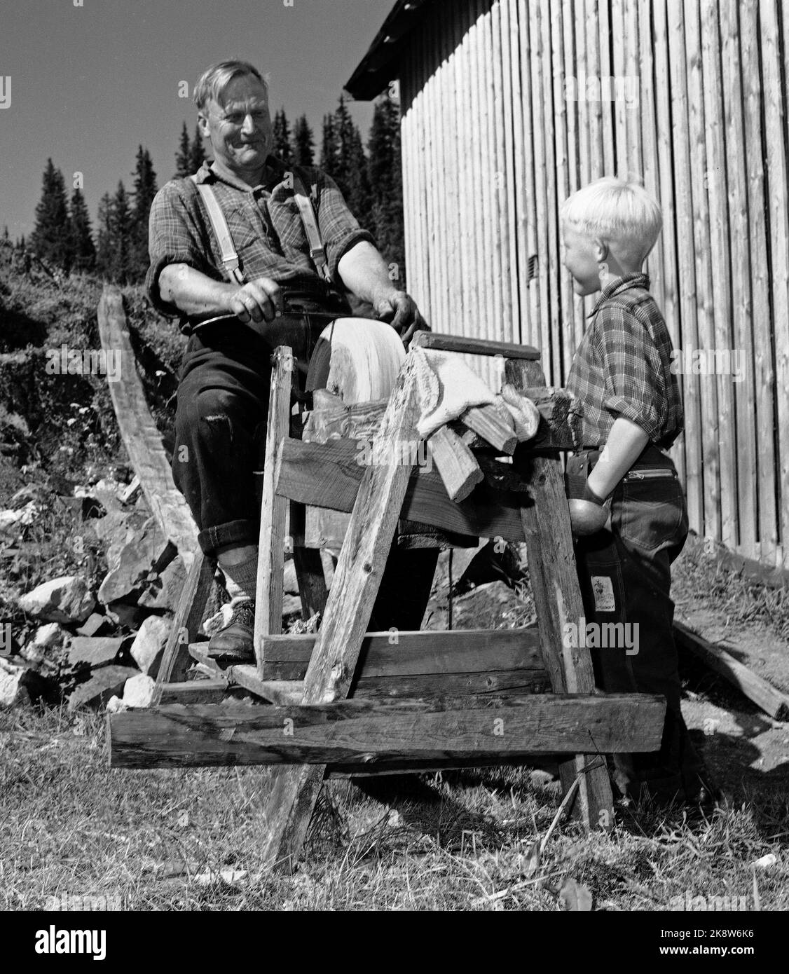 Flatdal in Telemark, August 1959. Fjellgården Ekre liegt 600 Meter über dem Meeresspiegel und in einem derart unwegsamen Gelände, dass "niemand denken würde, dass jemand es könnte BU". Hier der Bauer selbst, Bjørn Ekre, der auf dem Schleifstein im Hof grinst, mit seinem Sohn Bjarne (7) als Zuschauer. So wird ein alter Griff von Vater zu Sohn vererbt. Foto: Aage Storløkken / Aktuell / NTB Stockfoto