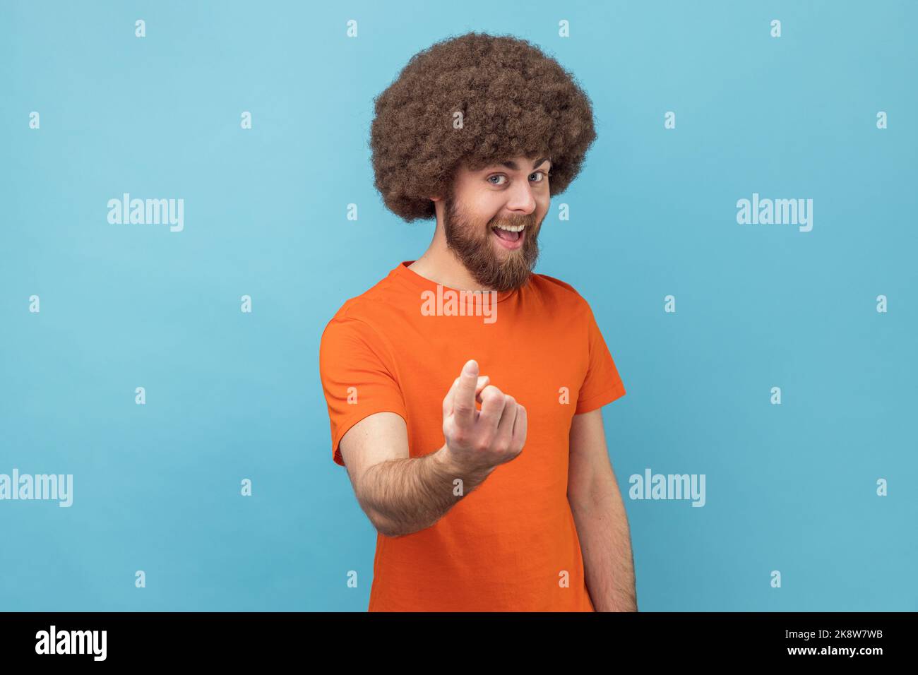 Kommen Sie hierher. Porträt eines zufriedenen lächelnden Mannes mit Afro-Frisur in orangefarbenem T-Shirt, der mit Fingergesten ruft und zu Kommen Sie herein. einlädt Innenaufnahme des Studios isoliert auf blauem Hintergrund. Stockfoto