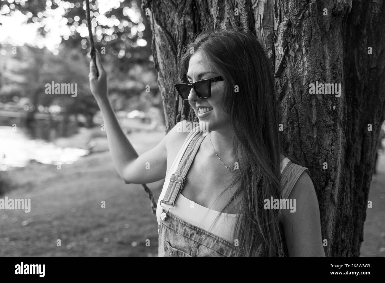 Schöne junge Frau mit Sonnenbrille, die sich auf einen Baum lehnt und zur Kamera schaut und in einem Park lächelt Stockfoto