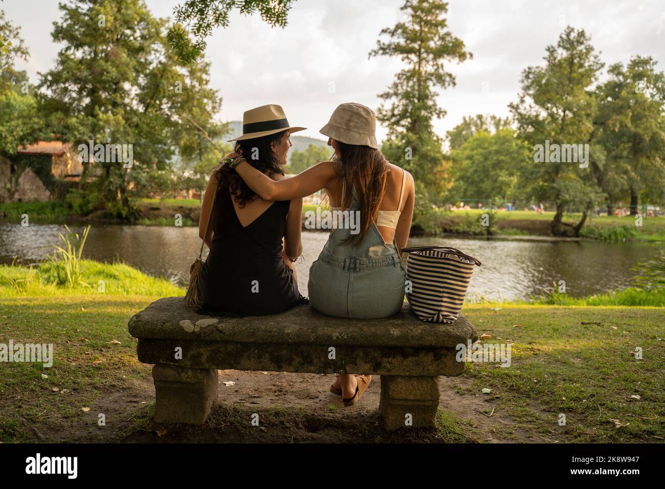 Zwei Freundinnen mit Hüten, die auf einer Steinbank vor dem Fluss sitzen und sich unterhalten Stockfoto