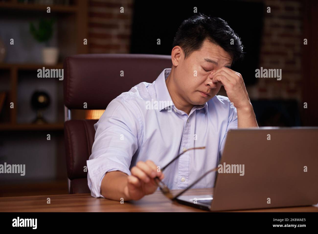 Deprimierter asiatischer Mann sitzt im Büro. Massiert den Nasenrücken vor Ermüdung der Augen. Schlechte Sicht nach dem Bildschirm. Stress, Müdigkeit, Burnout Stockfoto