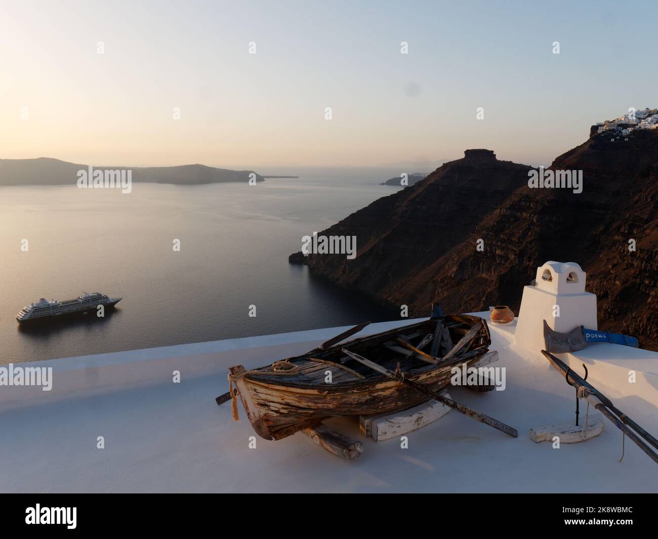 Blick auf die Caldera bei Sonnenuntergang über Firostefani mit Skaros Rock rechts. Griechische Kykladen-Insel Santorin in der Ägäis. Holzboot im Vordergrund auf dem Dach Stockfoto