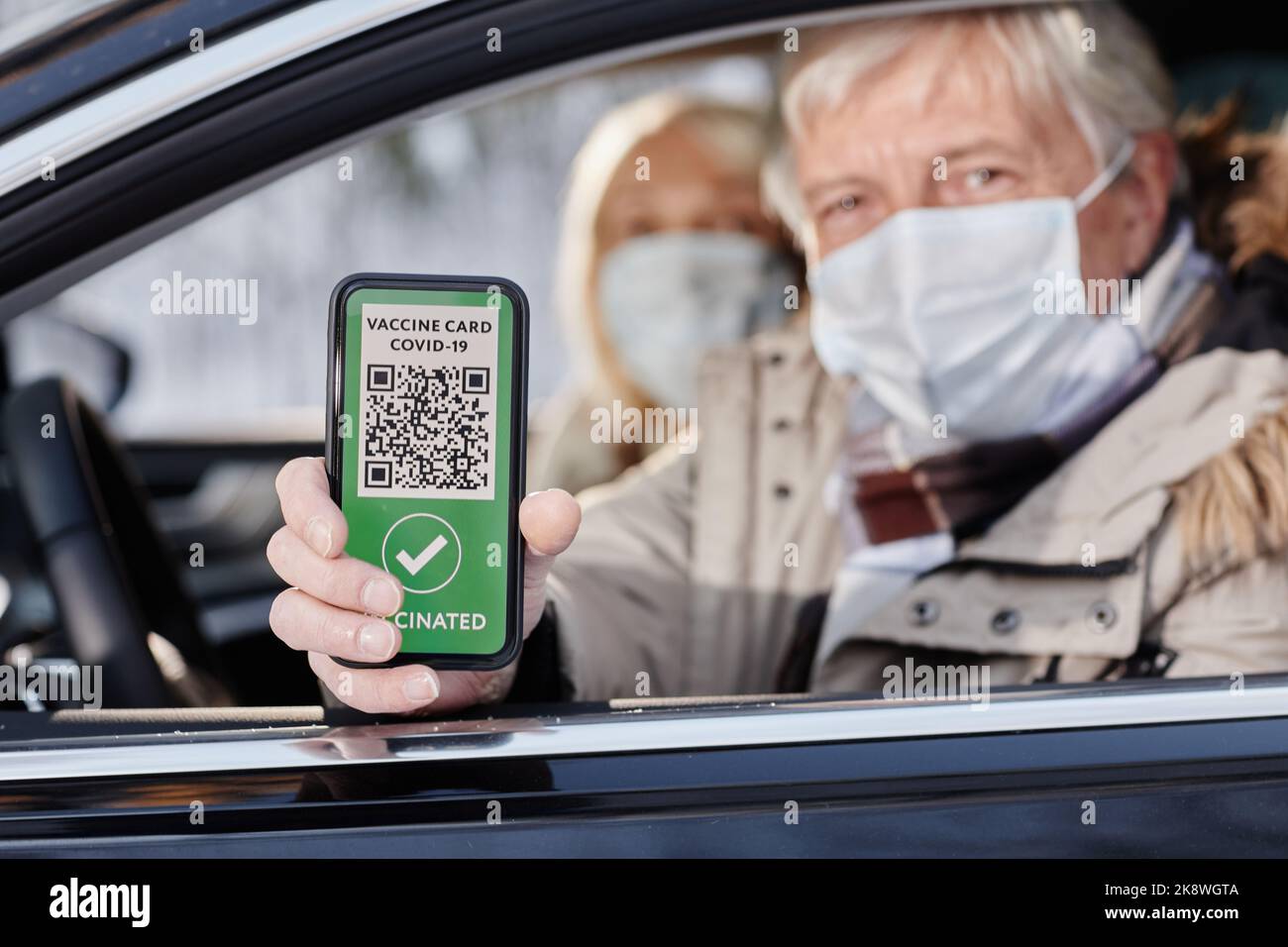 Älterer Mann, der den digitalen Impfcode vom Autofenster aus zeigt und während der Winterferien eine Maske trägt, Platz zum Kopieren Stockfoto