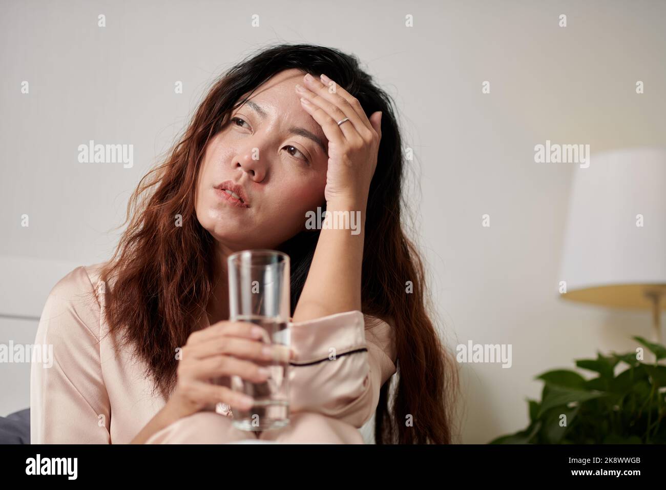 Kranke Frau, die an Kopfschmerzen und Fieber leidet, trinkt ein Glas Wasser und berührt die Stirn Stockfoto