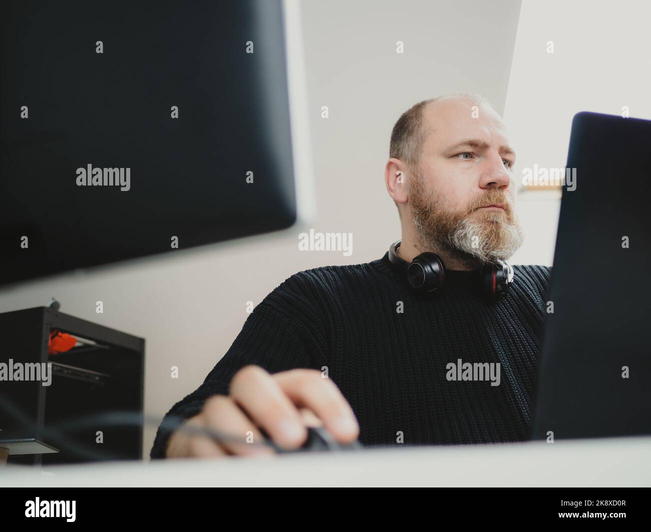 Erwachsener, bärtiger Mann, der an einem Laptop arbeitet und zwei Monitore von zu Hause aus verwendet Stockfoto