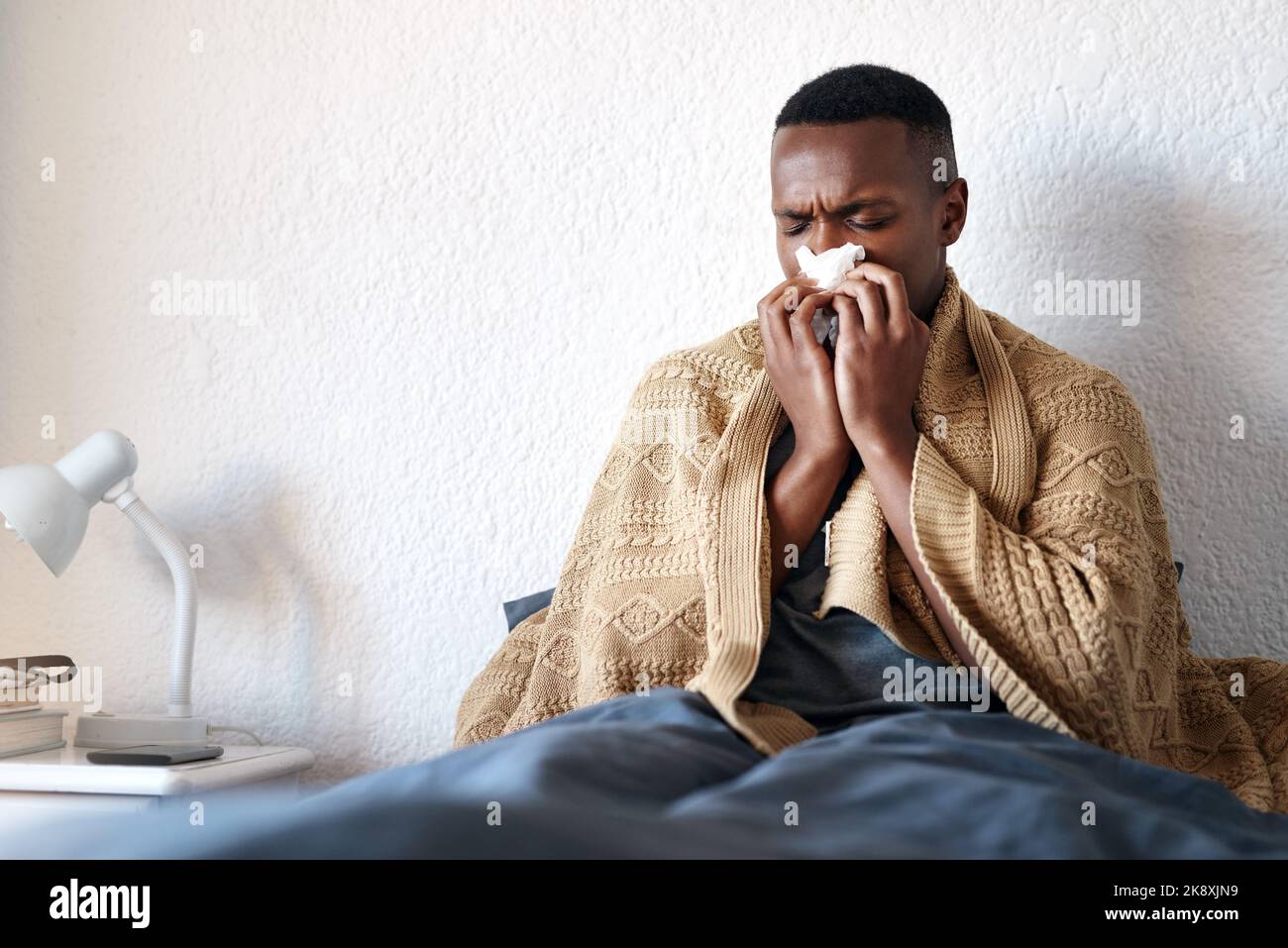 Ein hübscher junger Mann sitzt in seinem Bett und bläst seine Nase, während er an einer Erkältung leidet. Stockfoto