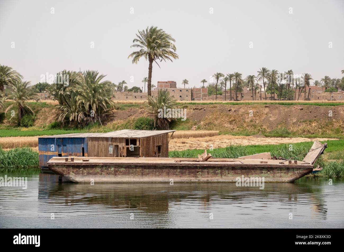 Ein Stahlkahn, der an den Ufern des Nils mit Palmen und Häusern in der Ferne vertäut ist Stockfoto