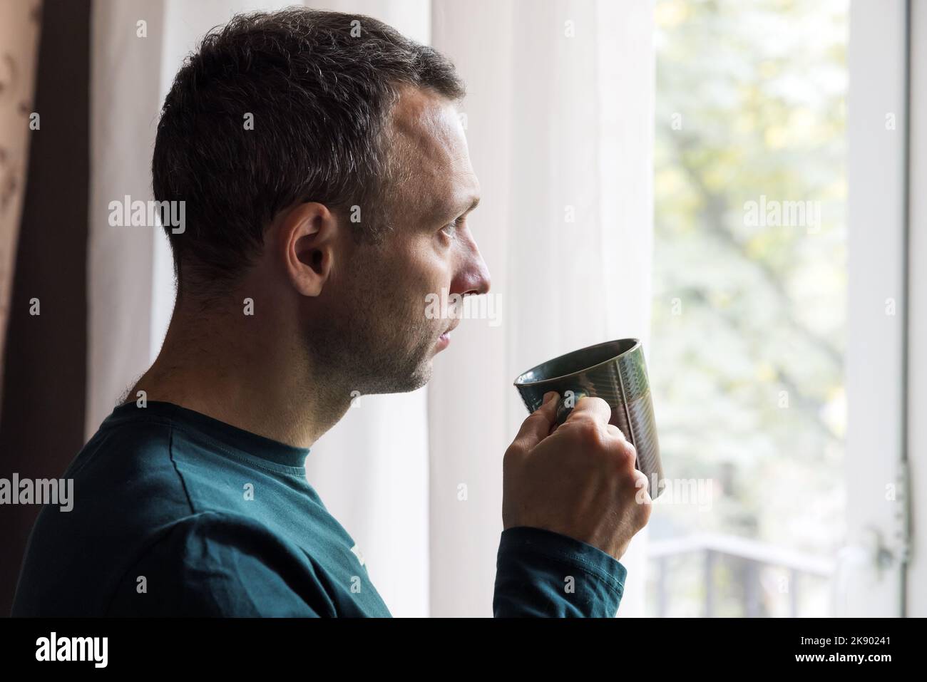 Profilporträt eines jungen erwachsenen kaukasischen Mannes, der vor einem hellen Fenster mit einer Tasse Kaffee in der Hand steht Stockfoto