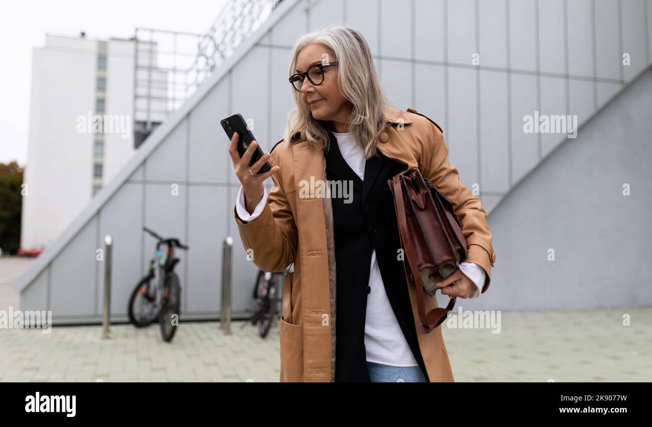 Ältere Erwachsene grauhaarige Geschäftsfrau eilt zu einem Treffen und schaut auf ein Mobiltelefon, 50-jährige europäische Frau, Unternehmertum Entwicklung und Stockfoto