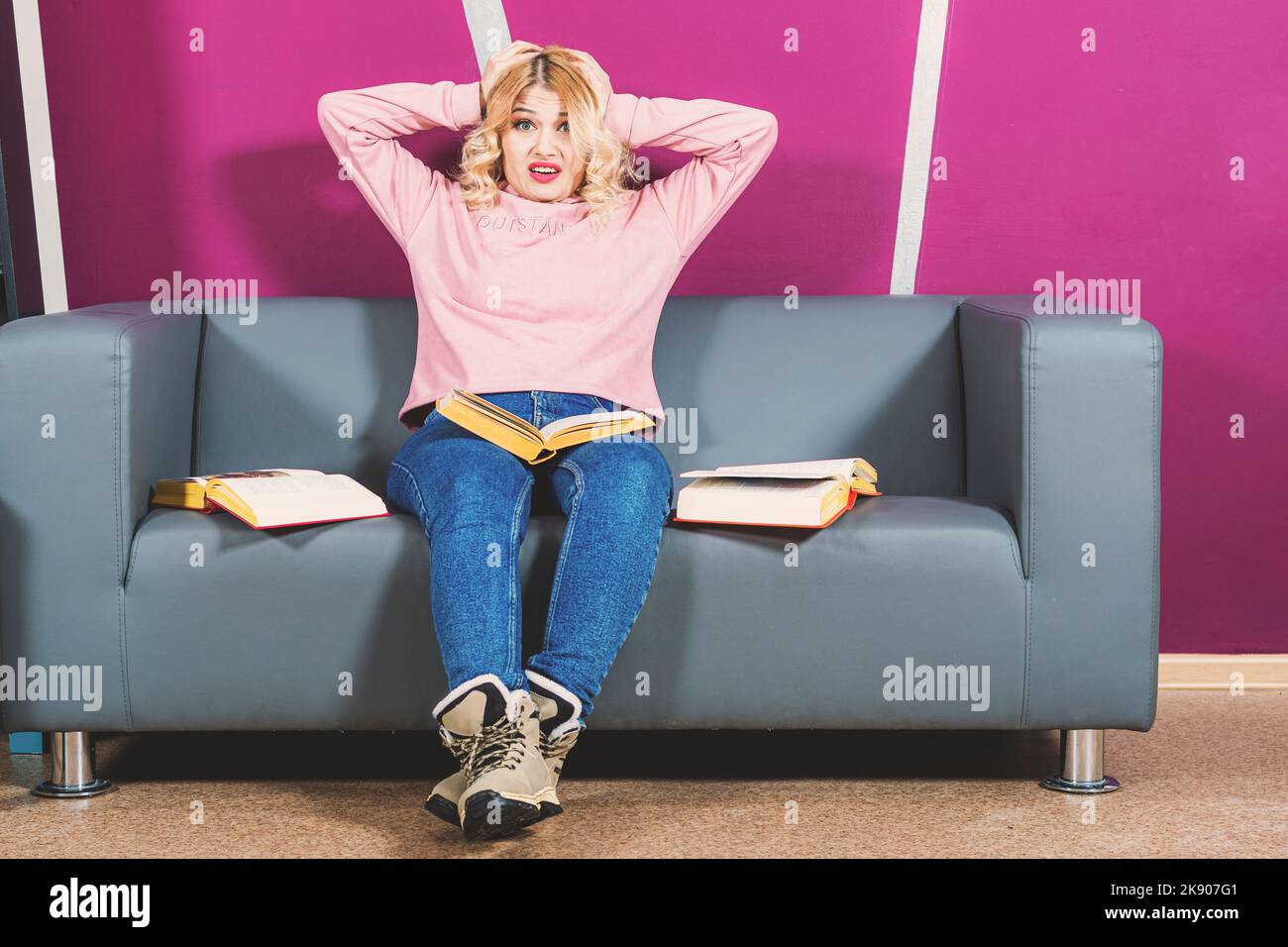 Das junge blonde Mädchen sitzt auf dem Sofa im Zimmer und hält den Kopf mit den Händen. Angesichts der Emotionen der Panik. Um sie herum sind offene Bücher. Lernschwierigkeiten. Stockfoto