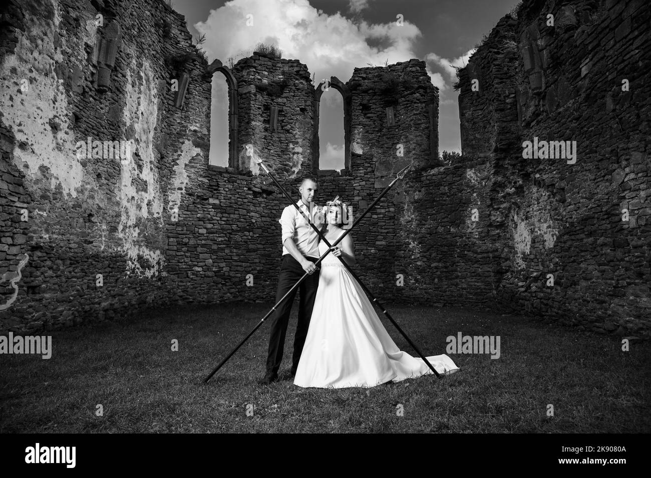 Eine Schwarz-Weiß-Aufnahme von einem schönen Paar in Anzug und Hochzeitskleid posiert vor der Kamera mit Speeren in einem alten Schloss Stockfoto