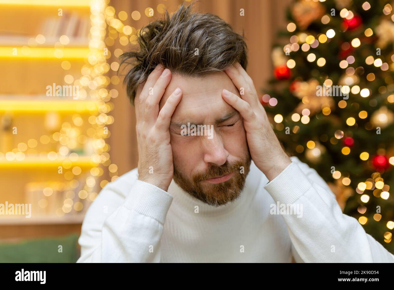 Ein Mann hat zu Weihnachten Kopfschmerzen, ein Nahaufnahme-Foto am Silvesterabend in der Nähe des Baumes, ein Mann hält seinen Kopf mit den Händen, krank, sitzt allein zu Hause auf dem Sofa. Stockfoto