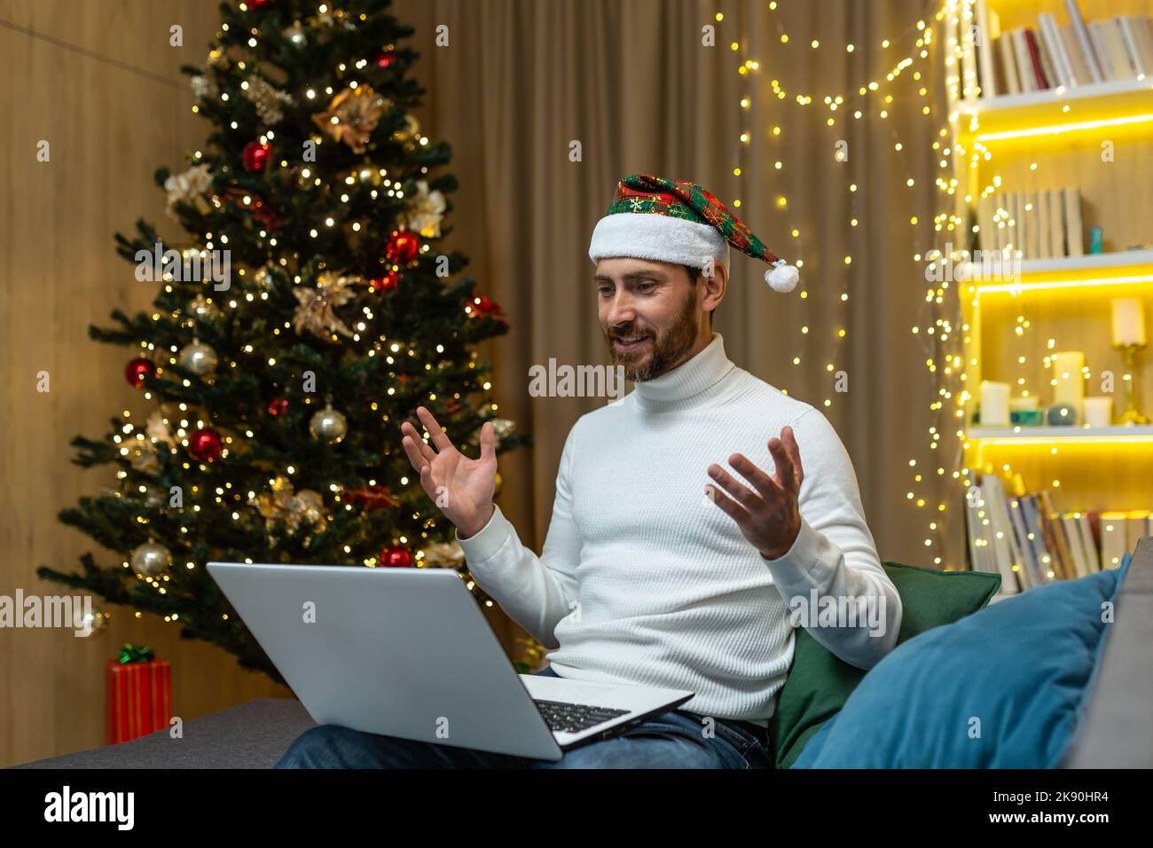 Mann feiert weihnachten und Neujahr allein zu Hause, mit Laptop für Videoanruf und Fernkommunikation mit Freunden online, sitzt auf dem Sofa im Wohnzimmer in der Nähe von Baum, glücklich in Hut. Stockfoto