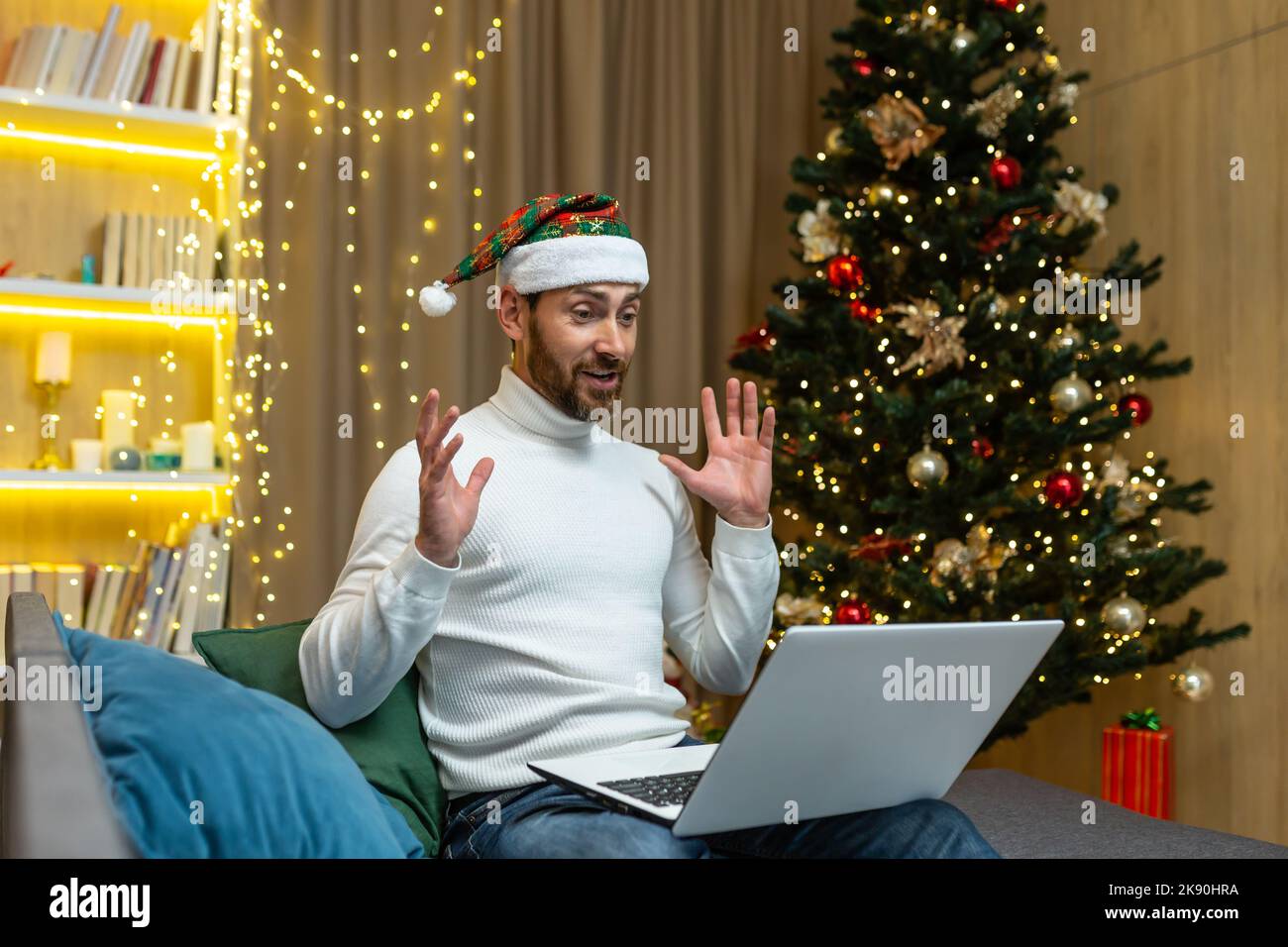 Mann feiert weihnachten und Neujahr allein zu Hause, mit Laptop für Videoanruf und Fernkommunikation mit Freunden online, sitzt auf dem Sofa im Wohnzimmer in der Nähe von Baum, glücklich in Hut. Stockfoto