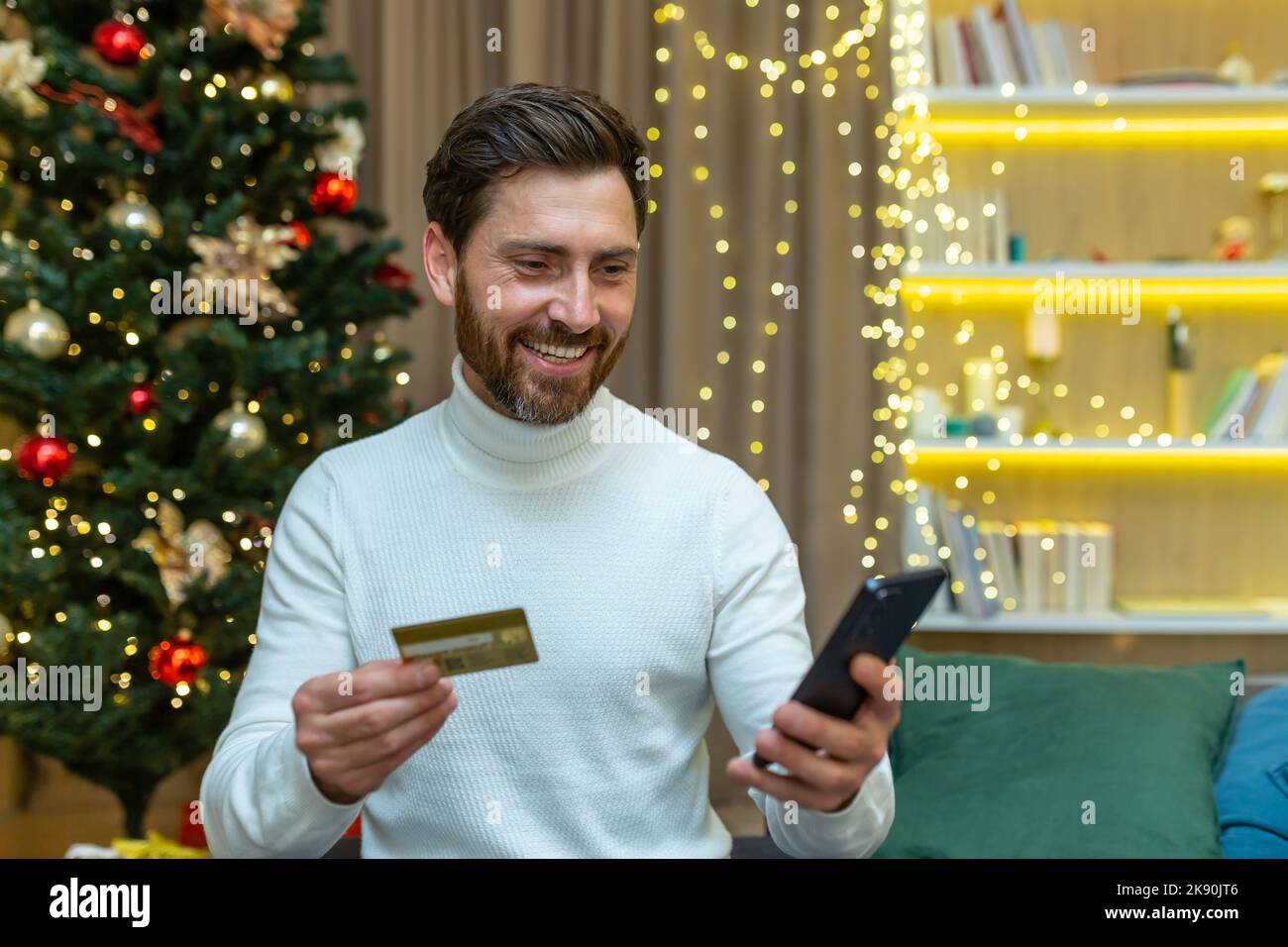 Mann allein zu Weihnachten zu Hause feiert Neujahr, sitzt auf dem Sofa und hält Smartphone und Bankkreditkarte, glücklich lächelnd Online-Shopping Auswahl von Geschenken für Freunde und Familie aus der Ferne. Stockfoto