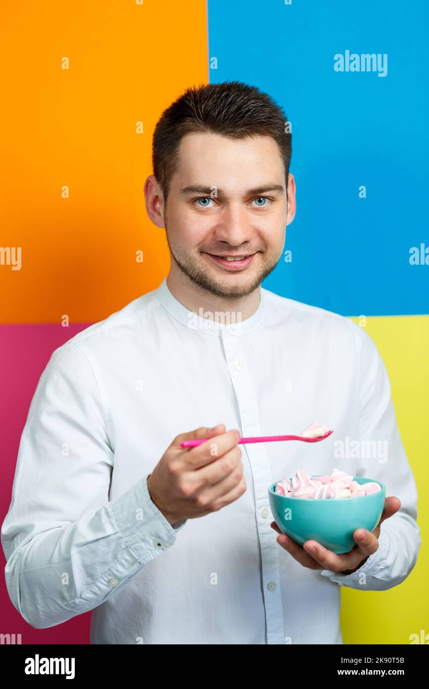 Ein Mann auf gelbem, blauem, orangefarbenem, rosa Hintergrund in einem weißen Hemd mit einem türkisfarbenen Teller Marshmallows und einem rosa Löffel. Der Typ mit einem Lächeln. Ein im Stockfoto