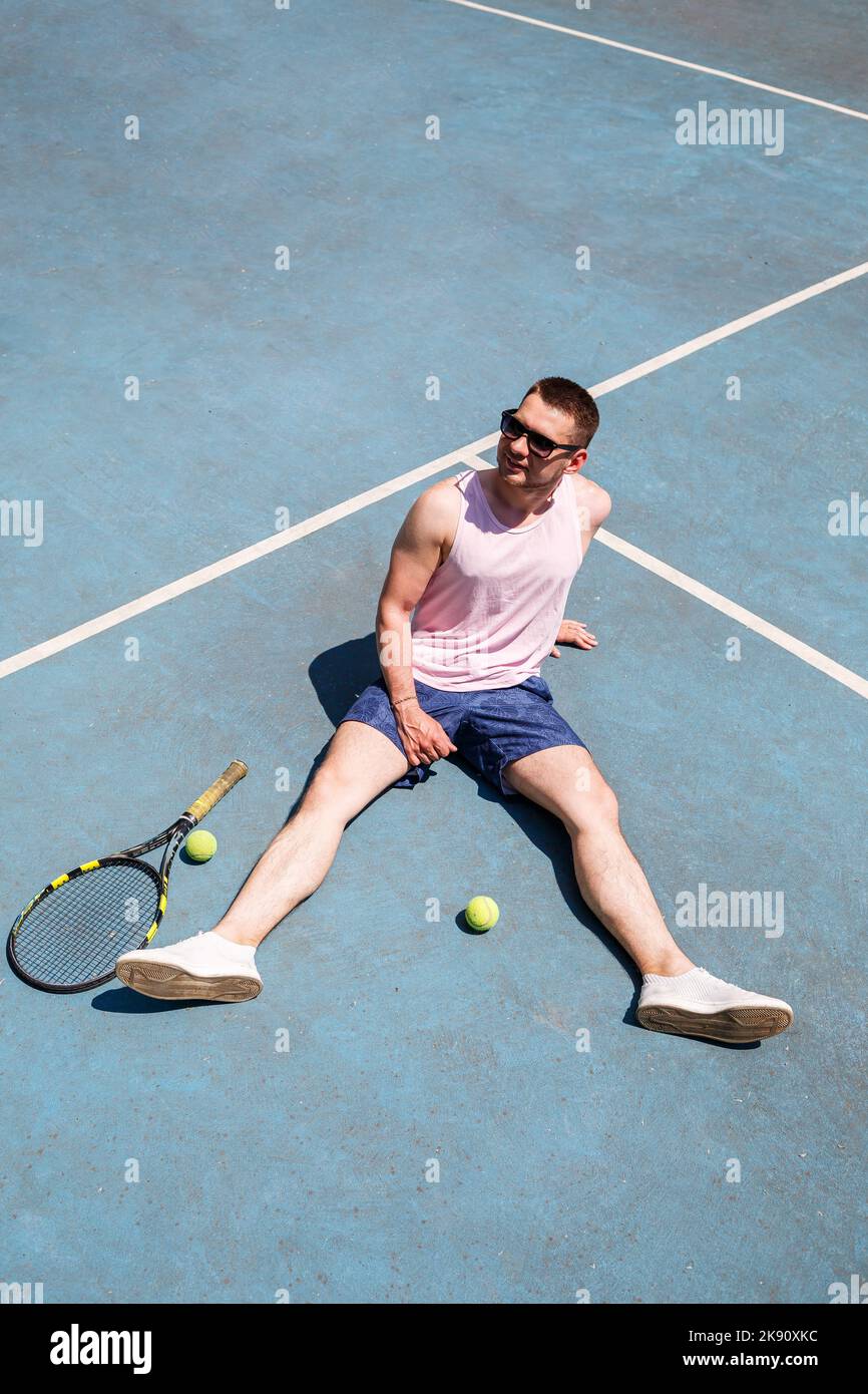 Ein hübscher Mann in einem T-Shirt und Shorts sitzt mit einem Schläger auf einem Tennisplatz. Mann in Sonnenbrille Stockfoto