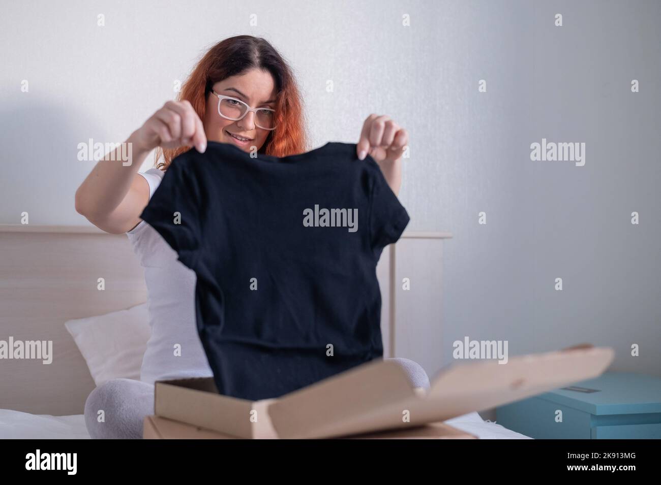Frau packt online ein T-Shirt aus, während sie im Bett sitzt. Lieferung nach Hause Stockfoto
