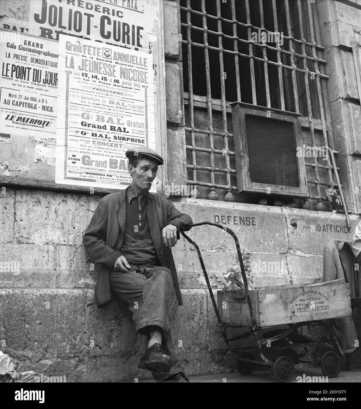 Frankreich im Jahr 1950s. Ein Mann auf der Straße sitzt mit einer Zigarette. Er lehnt sich auf einen primitiven Wagen, an der Wand hinter ihm sind Plakatwände für frühere und aufwühlende Ereignisse sichtbar. Cannes Frankreich 1950 Kristoffersson Ref. AZ60-11 Stockfoto