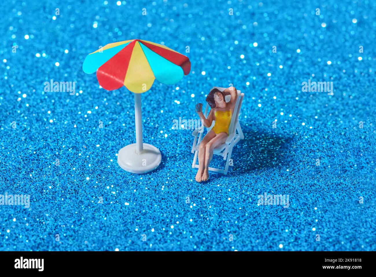 Hoher Winkel von Miniatur-Kunststoff-Dame auf Stuhl unter hellen bunten Regenschirm auf blau glitzernden Oberfläche sitzen Stockfoto