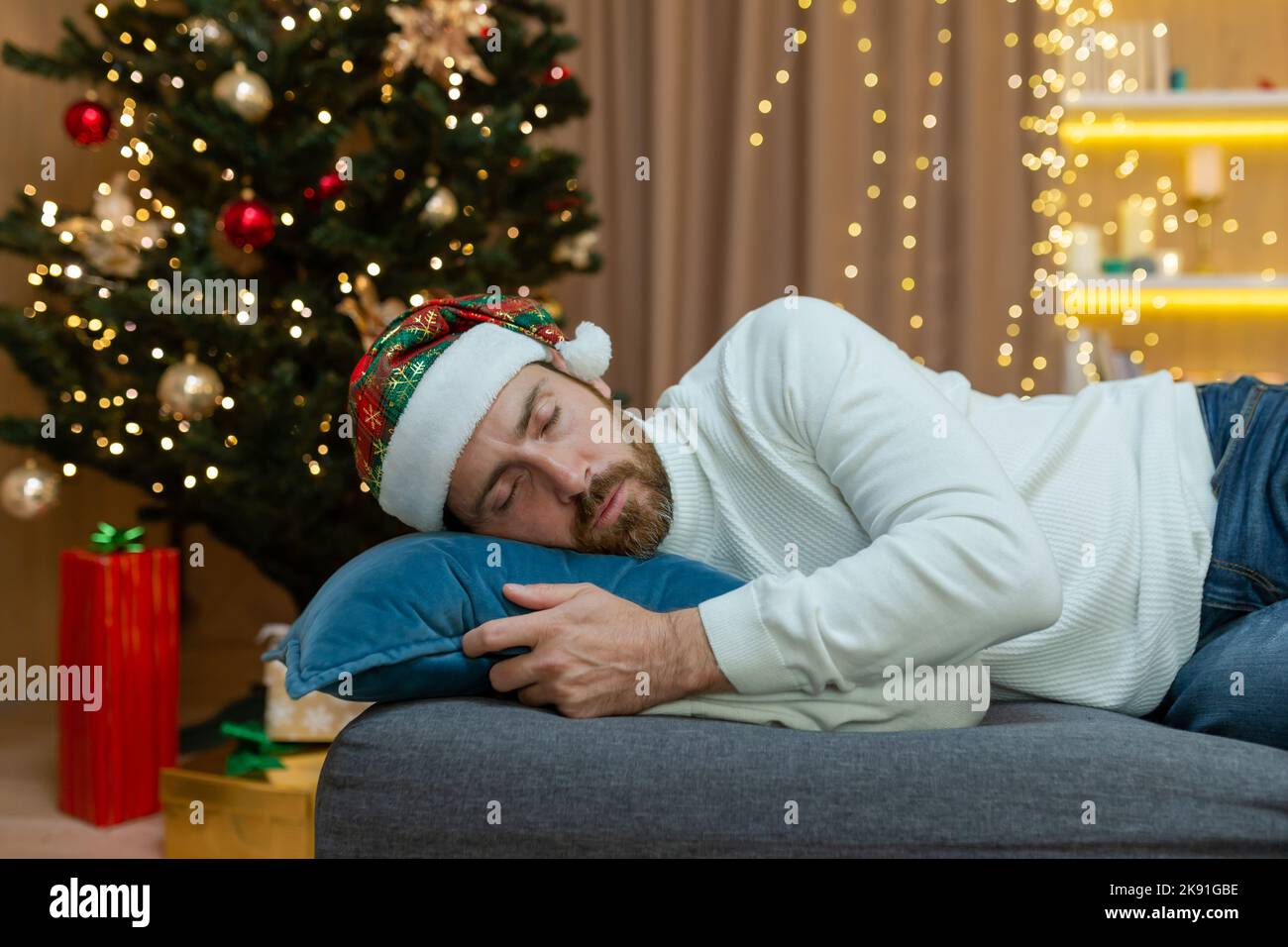 Ein Mann ist für Weihnachten müde, trägt einen Neujahrshut, schläft in der Nähe des Weihnachtsbaums, sitzt zu Hause im Wohnzimmer auf dem Sofa, nachdem er Weihnachten gefeiert hat. Stockfoto