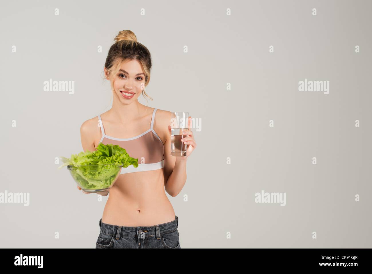 Lächelnde Frau mit schlankem Körper, die frischen Salat und ein Glas reines Wasser auf grau isoliert hält Stockfoto