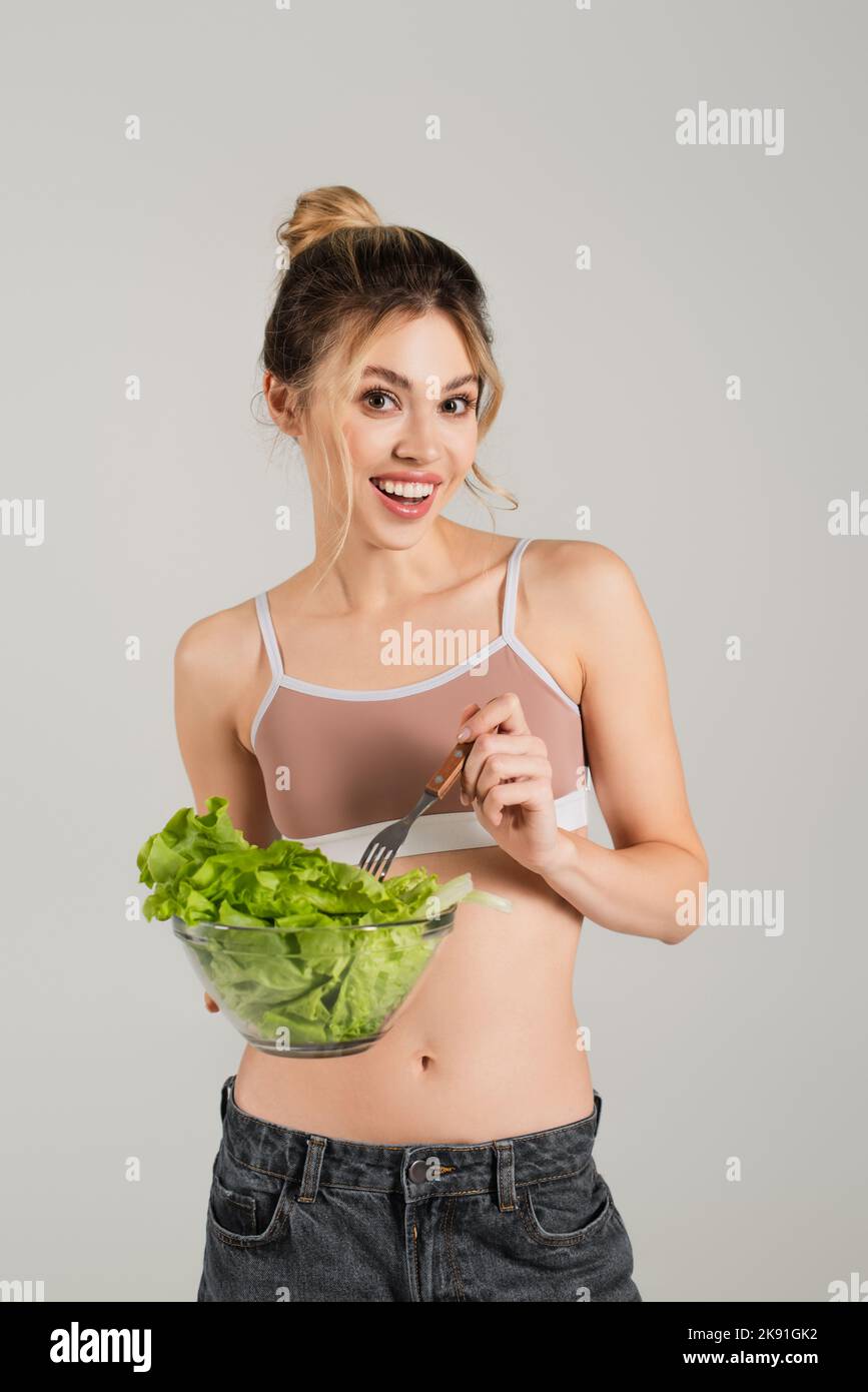 Fröhliche Frau in Sport Top hält Schüssel mit Salat und Gabel isoliert auf grau Stockfoto