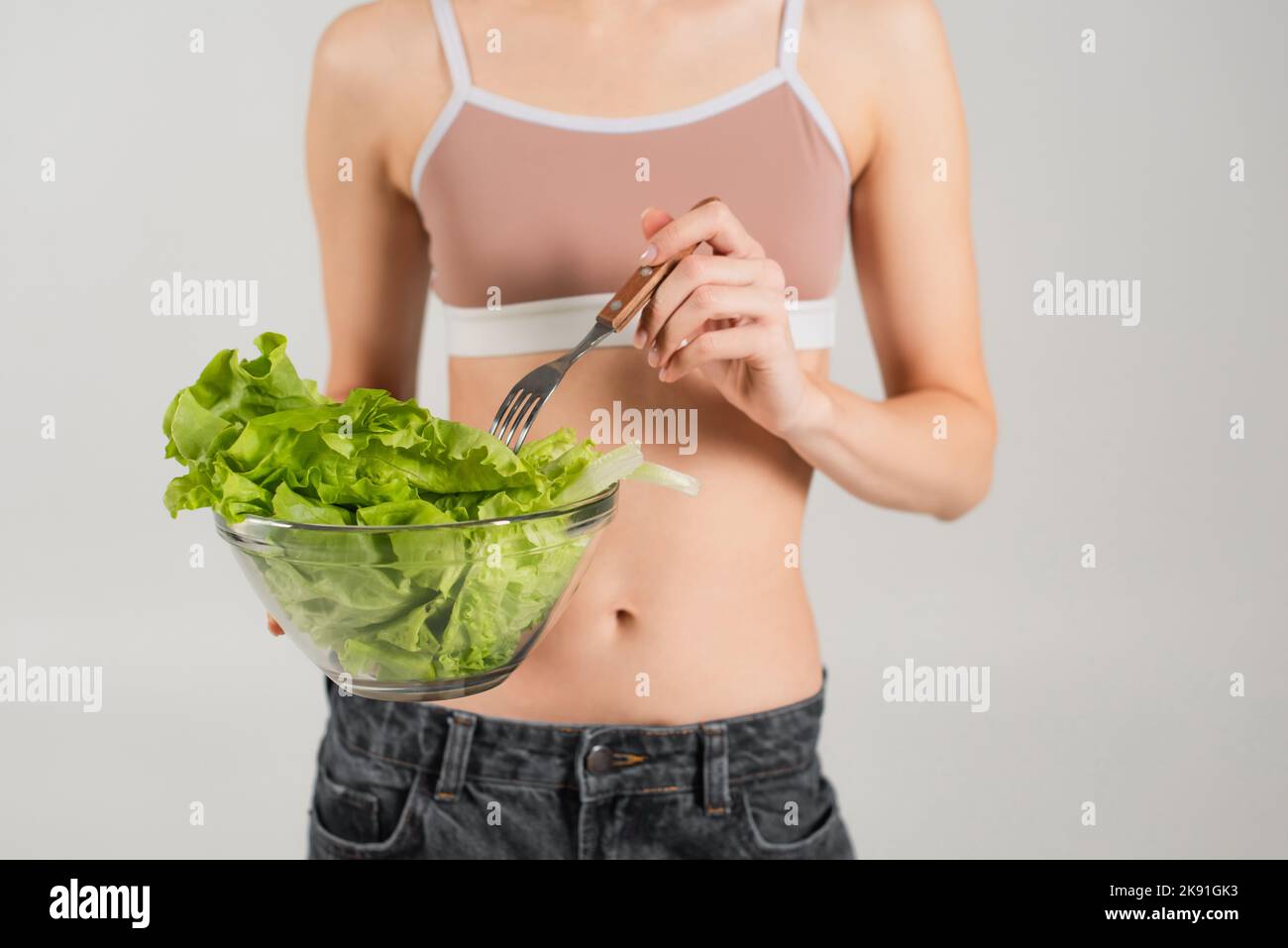 Beschnittene Ansicht einer Frau in einem Sporttop mit Gabel und frischem Salat, isoliert auf Grau Stockfoto