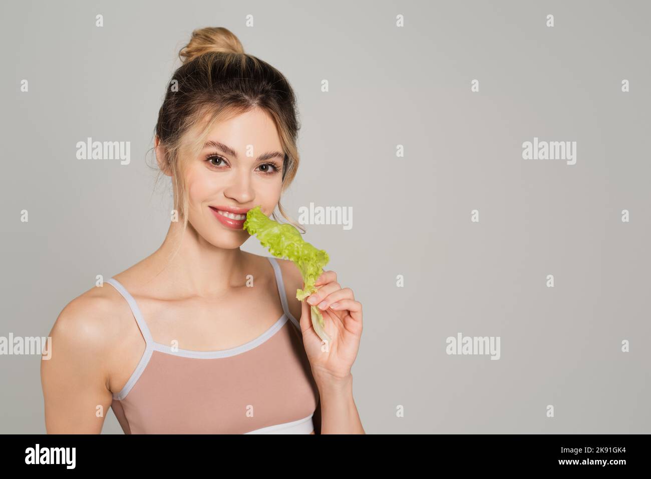 Hübsche Frau mit natürlichem Make-up, die frischen Salat hält und die Kamera isoliert auf Grau anschaut Stockfoto