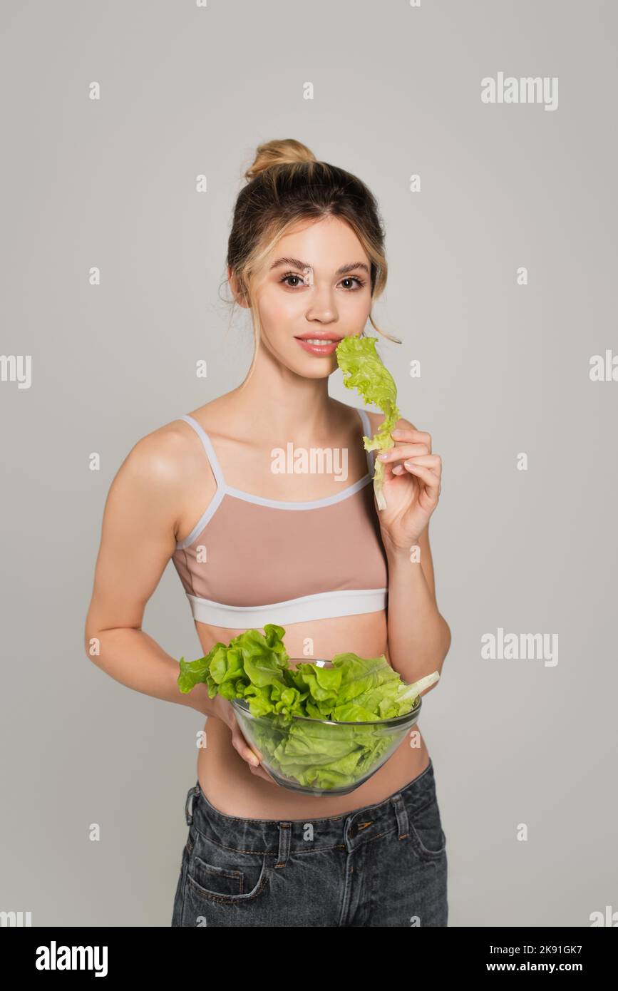 Lächelnde Frau mit schlankem Körper, der Salat hält und isoliert auf Grau auf die Kamera schaut Stockfoto