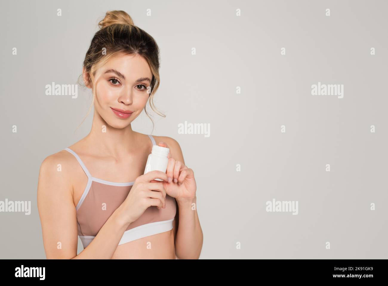 Hübsche Frau mit perfekter Haut, die Vitaminbehälter hält und die Kamera isoliert auf Grau anlächelt Stockfoto