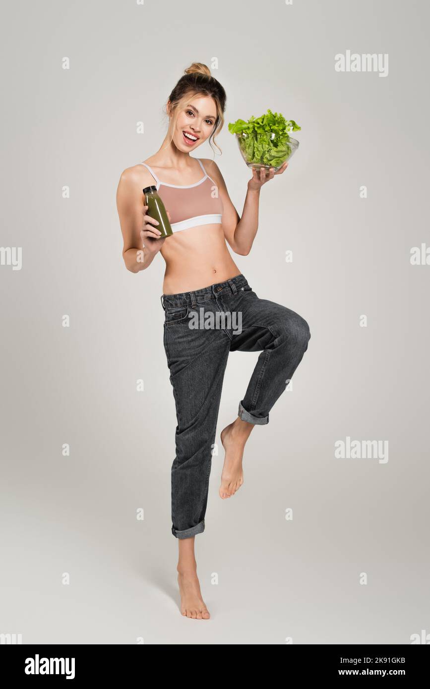 Aufgeregt und fit Frau posiert mit einer Flasche Smoothie und frischem Salat auf grauem Hintergrund Stockfoto