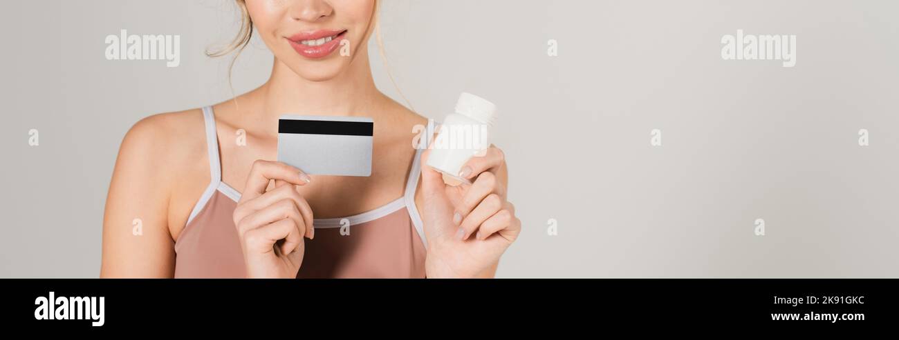 Teilansicht einer lächelnden Frau mit Kreditkarte und Vitaminen isoliert auf grauem Banner Stockfoto