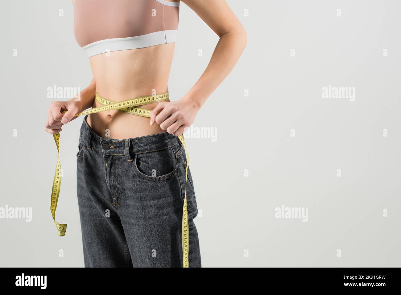 Cropped Ansicht der schlanken Frau in Sport-Top und Jeans Messung Taille isoliert auf grau Stockfoto