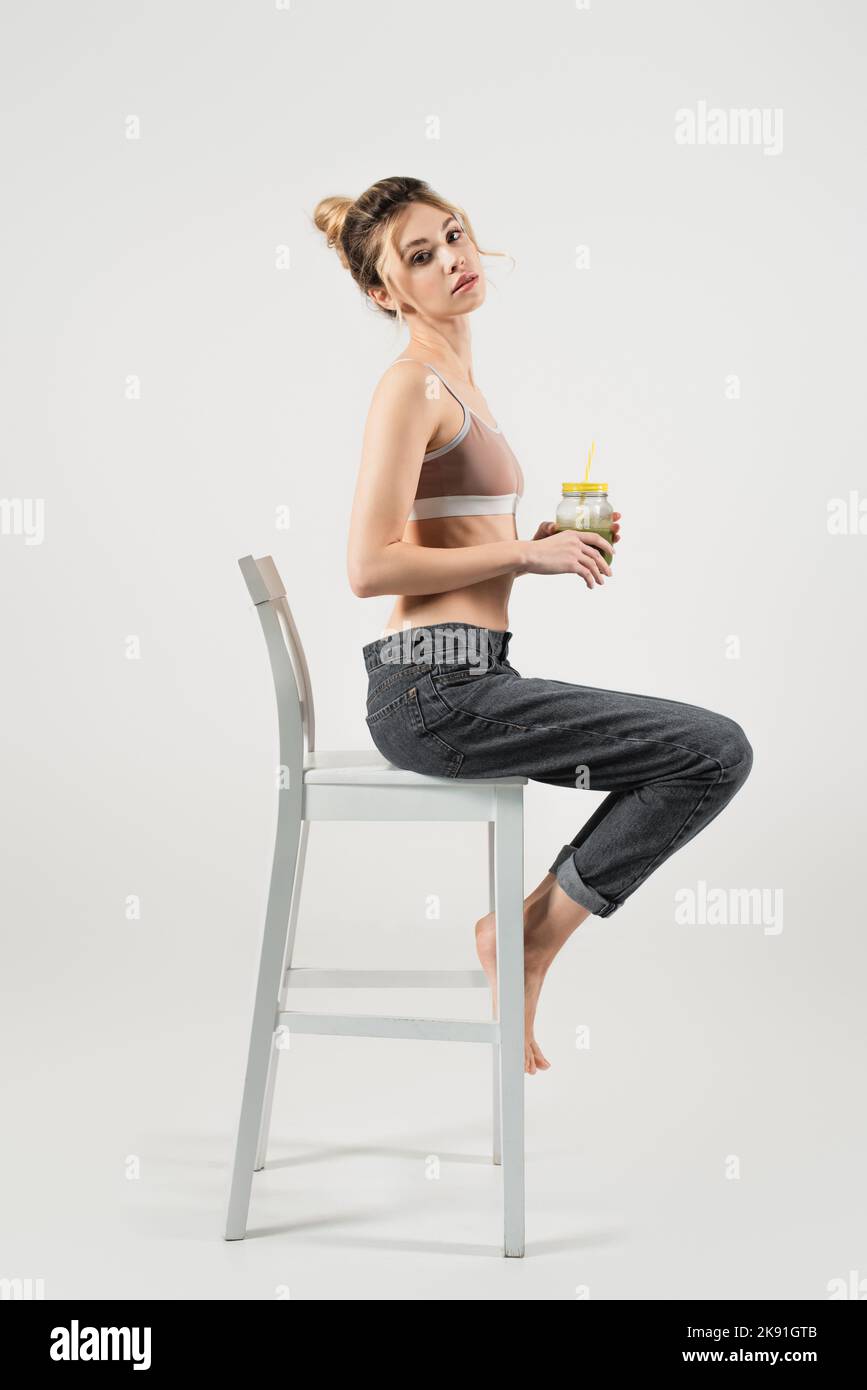 Schlanke Frau in Sporttop und Jeans hält Smoothie, während sie auf einem weißen Stuhl auf grauem Hintergrund sitzt Stockfoto