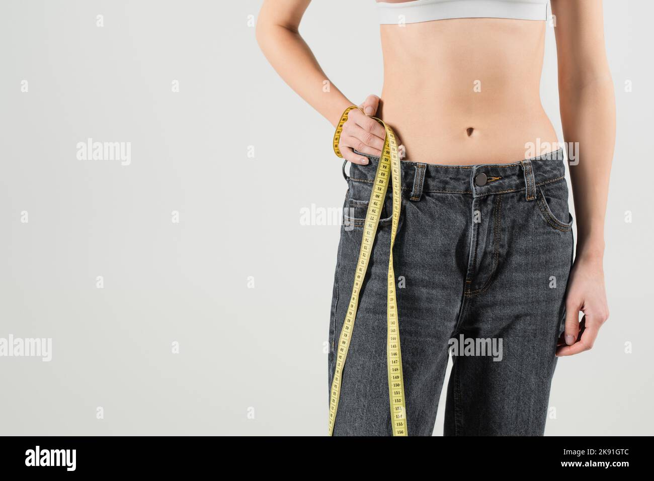 Verkürzte Ansicht einer fetten Frau in Jeans, die mit der Hand auf der Hüfte steht und ein Maßband auf Grau isoliert Stockfoto